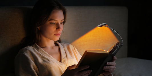 Woman reading comfortably in bed using a wide-beam amber LED clip-on book light to reduce eye strain.