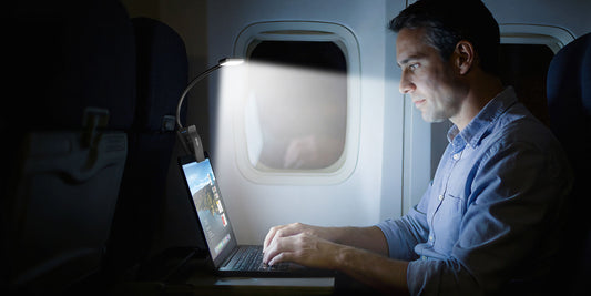 Man working on a laptop in a dark airplane cabin using a portable clip-on LED travel light.
