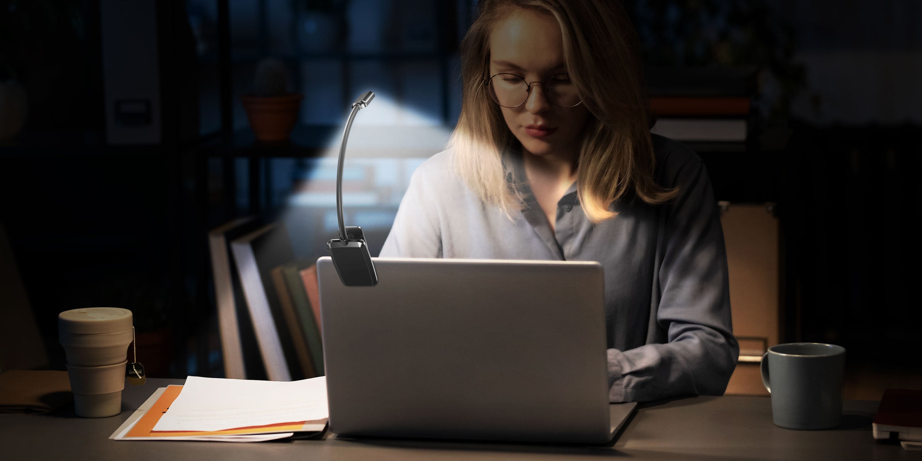 Woman working late at a desk with her laptop illuminated by a small clip-on LED white reading light.