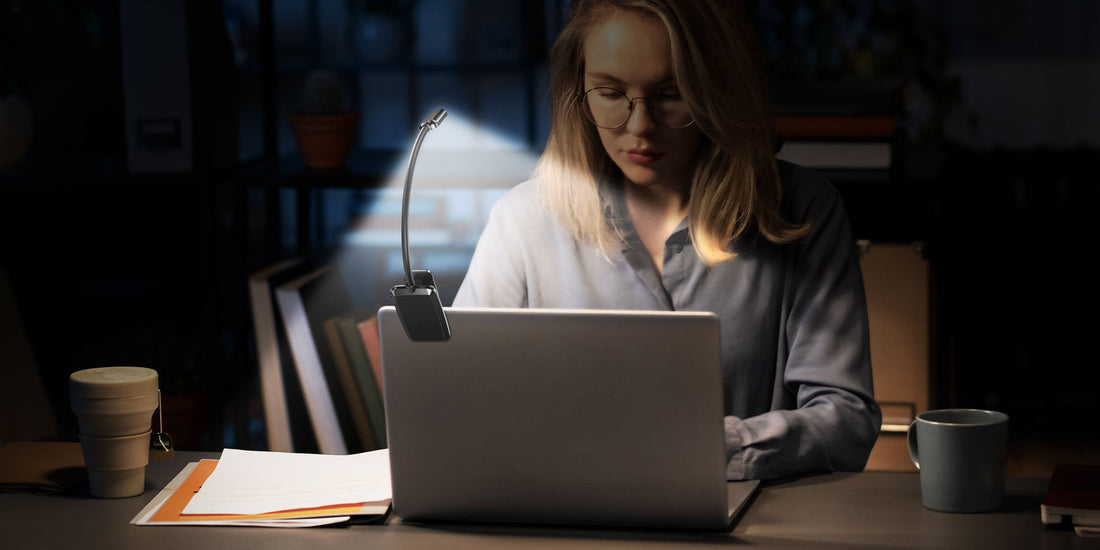 Woman working late at a desk with her laptop illuminated by a small clip-on LED white reading light.